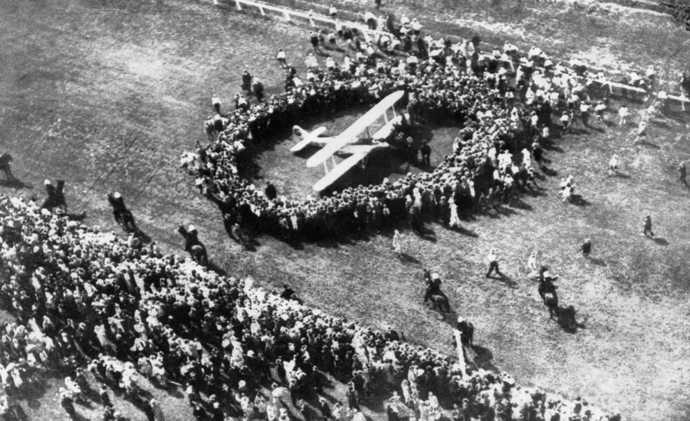 A crowd of people are gathered around a plane in the middle of a field.