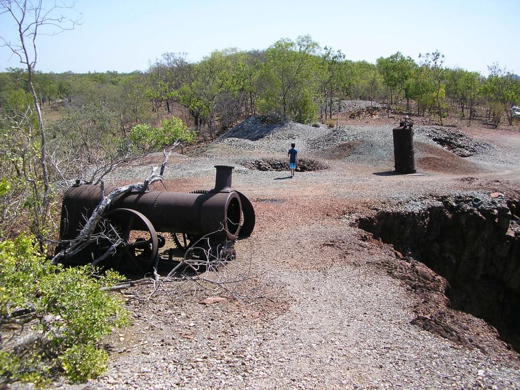 A man is standing in the middle of a dirt road next to a pile of pipes.