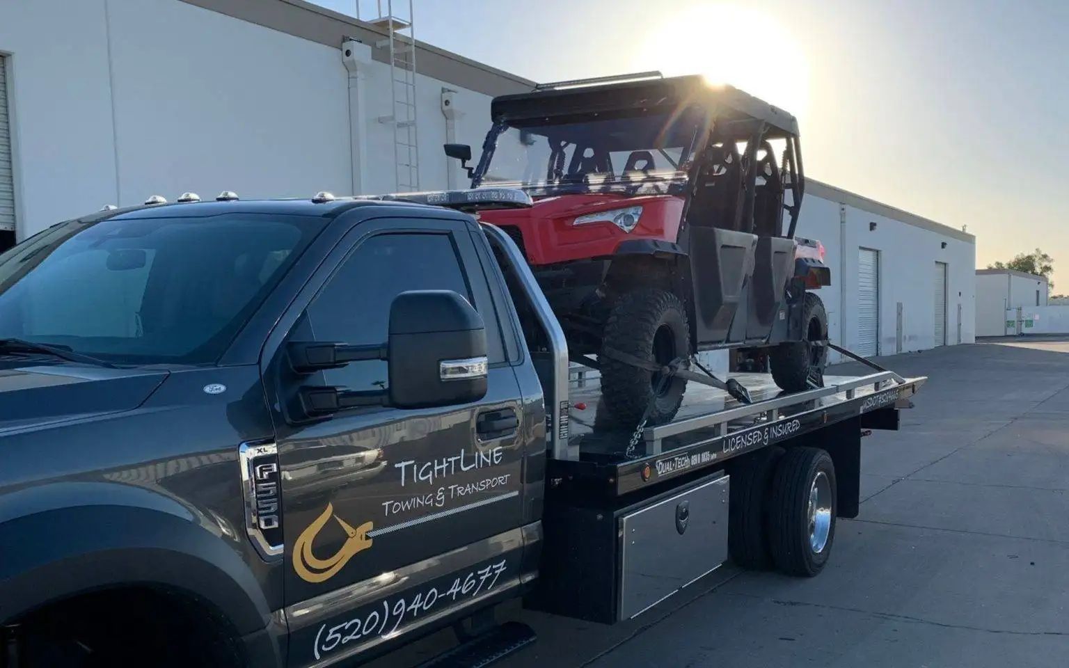 A flatbed tow truck carrying a red UTV with the sun shining in the background.