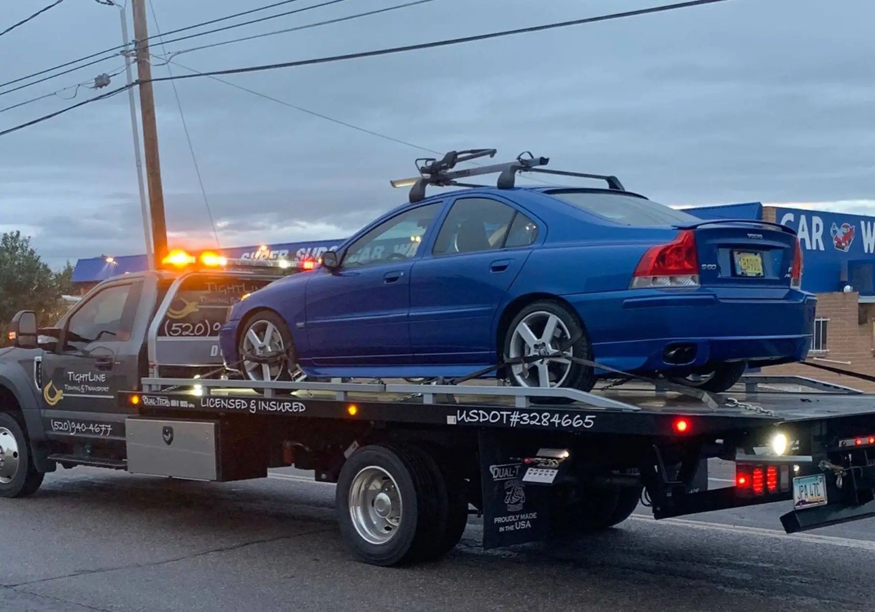 A blue car with a roof rack is towed on a flatbed truck in front of a car wash.