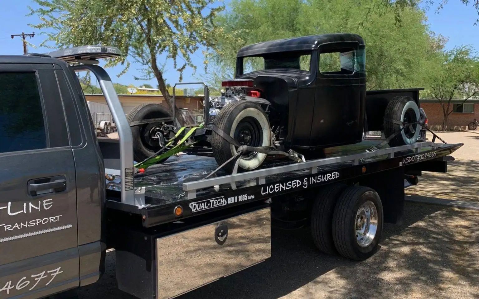 Black vintage truck on a flatbed tow truck. The truck has whitewall tires. Sunny outdoor setting.