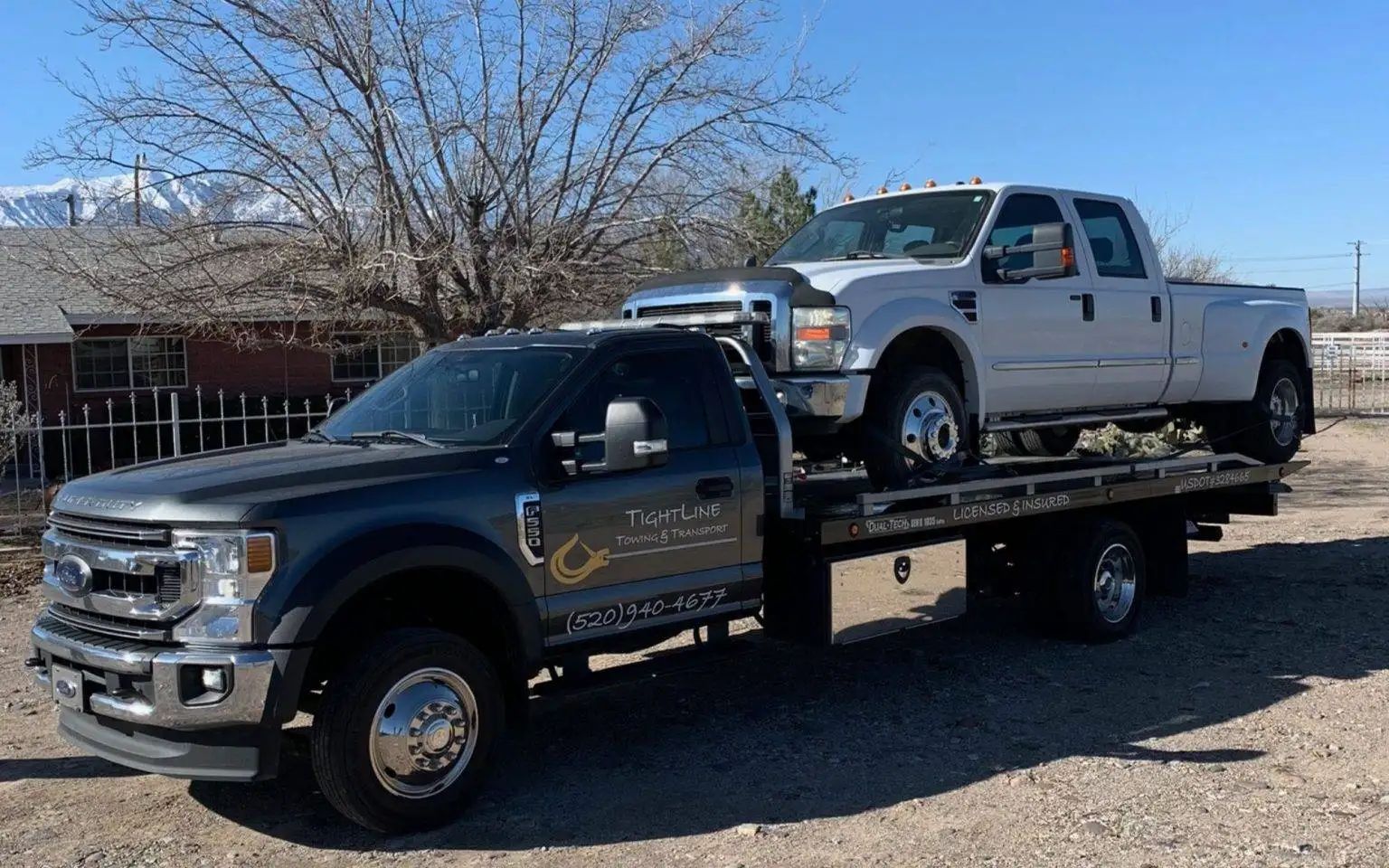Tow truck hauling a light-colored pickup truck. The vehicles are on a road with mountains in the background.
