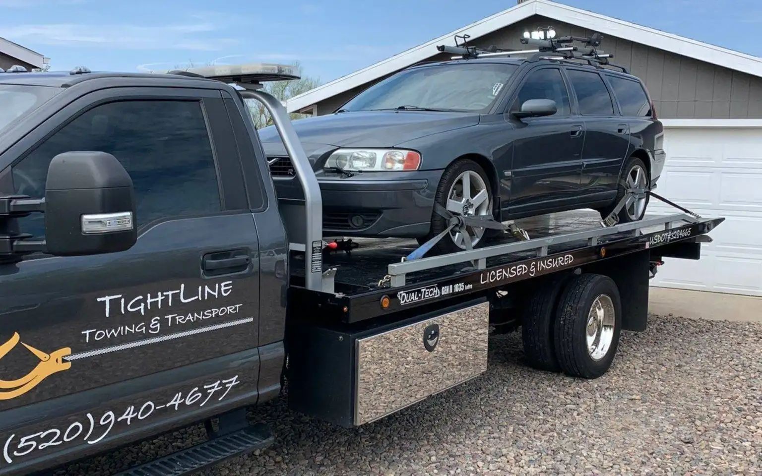 Grey Volvo station wagon on a flatbed tow truck, near a building with garage door; 