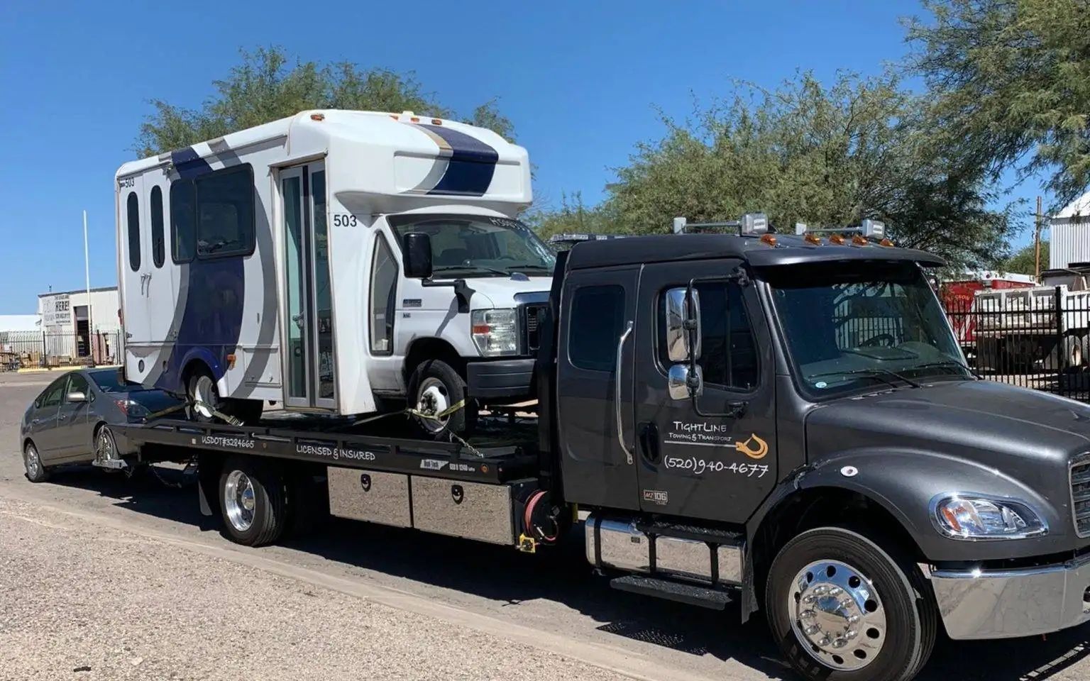 A disabled white and blue bus is towed by a dark grey tow truck on a sunny day.