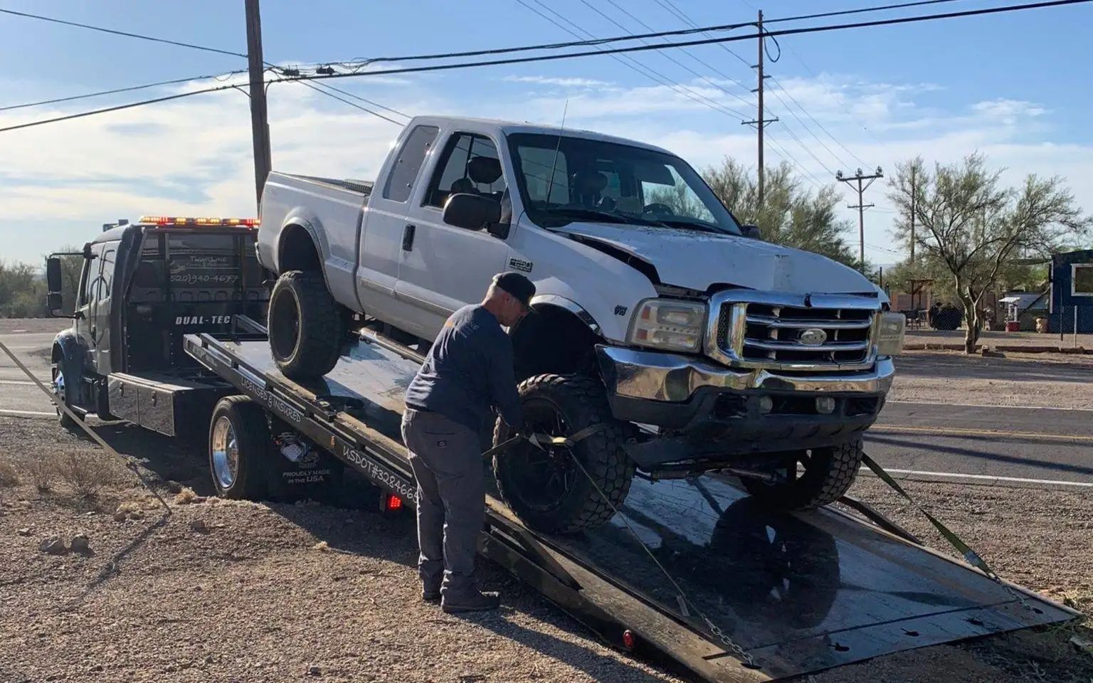 White truck on a tow truck being checked by a person on a sunny day near a road.