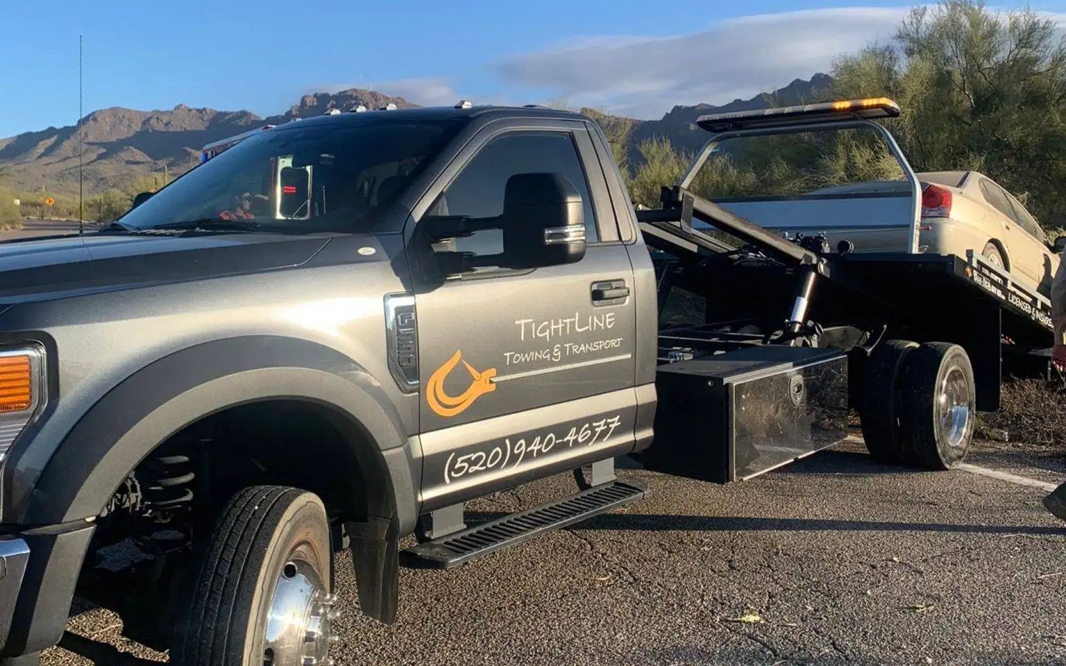 Gray tow truck with a light-colored car on its flatbed.  Mountains in the background.