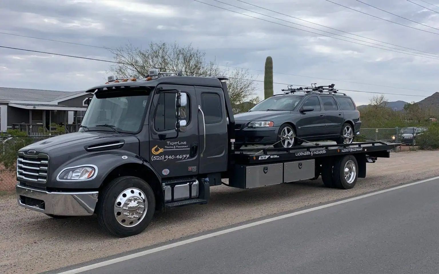 Dark tow truck with a gray car on its flatbed, roadside scene.
