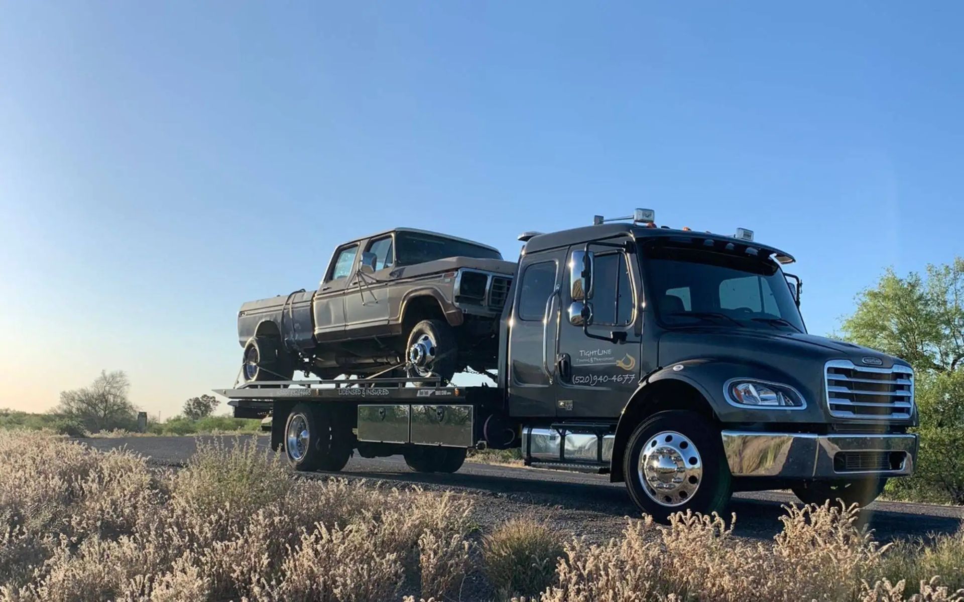 Tow truck carrying a rusty pickup truck on a sunny road.