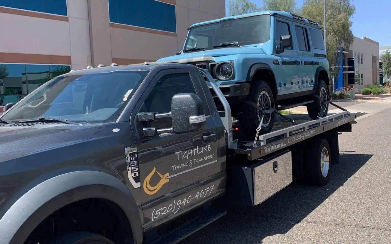 Blue SUV being towed on a flatbed truck. Both vehicles are on a street, in front of a building.