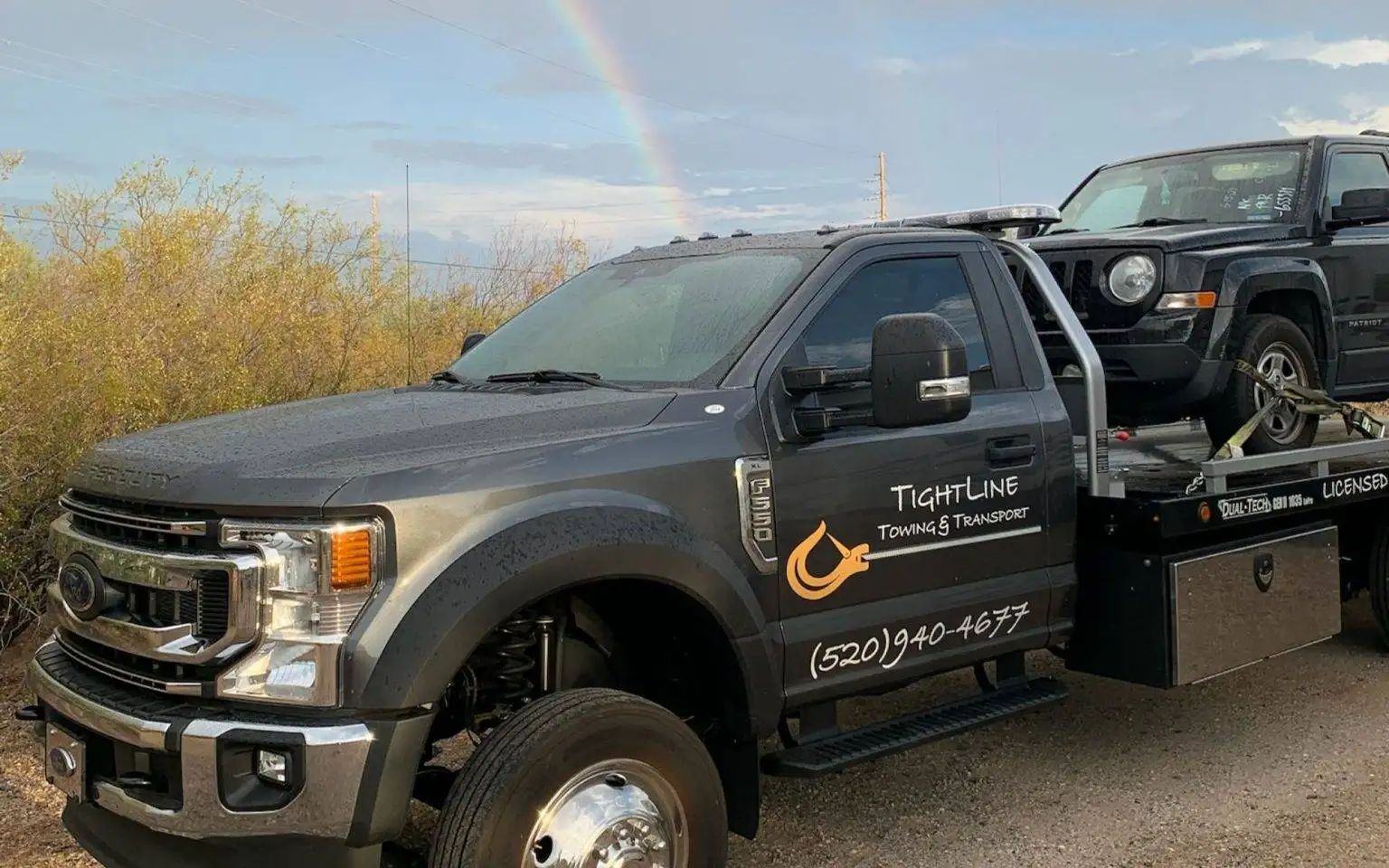 Tow truck with a black SUV on its flatbed, under a rainbow. Gray truck with logo on door, parked outdoors.