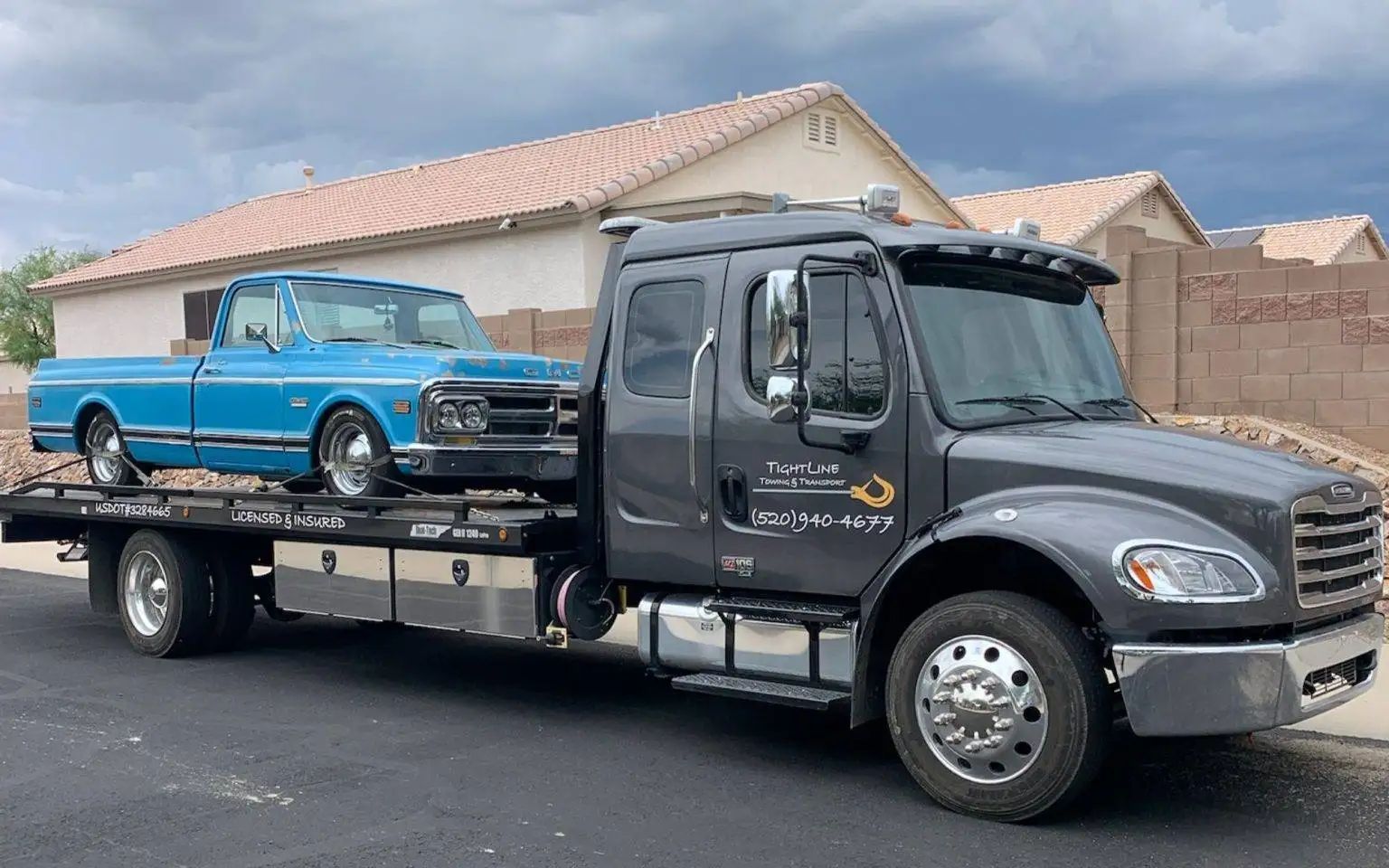 Blue classic truck on a gray tow truck in front of a house on a cloudy day.