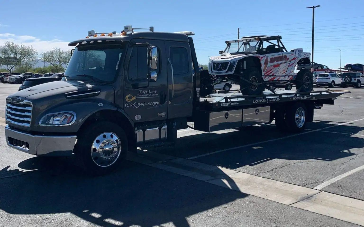 Gray tow truck with a white and red UTV loaded on the flatbed, parked outdoors.