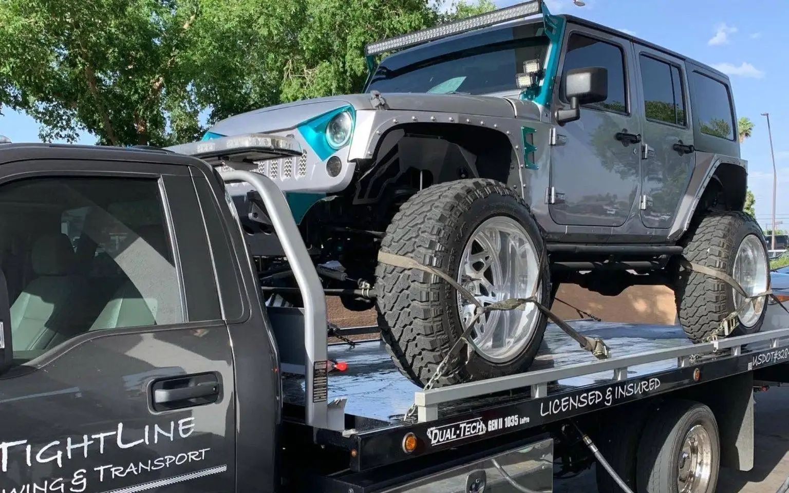 Silver Jeep with turquoise accents loaded on a tow truck, outdoors.