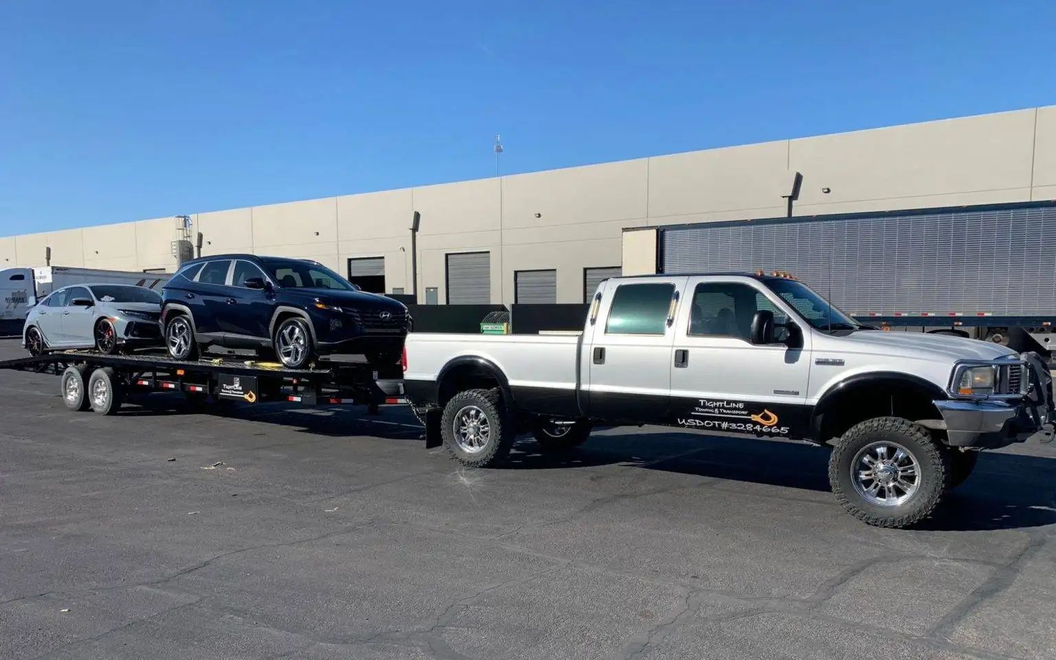 A lifted silver truck towing a trailer carrying two cars on a sunny day in front of a warehouse.