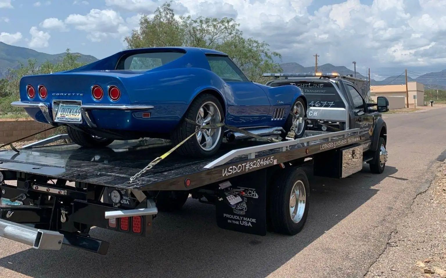 Blue Corvette sports car on a tow truck bed, in an outdoor setting with mountains.