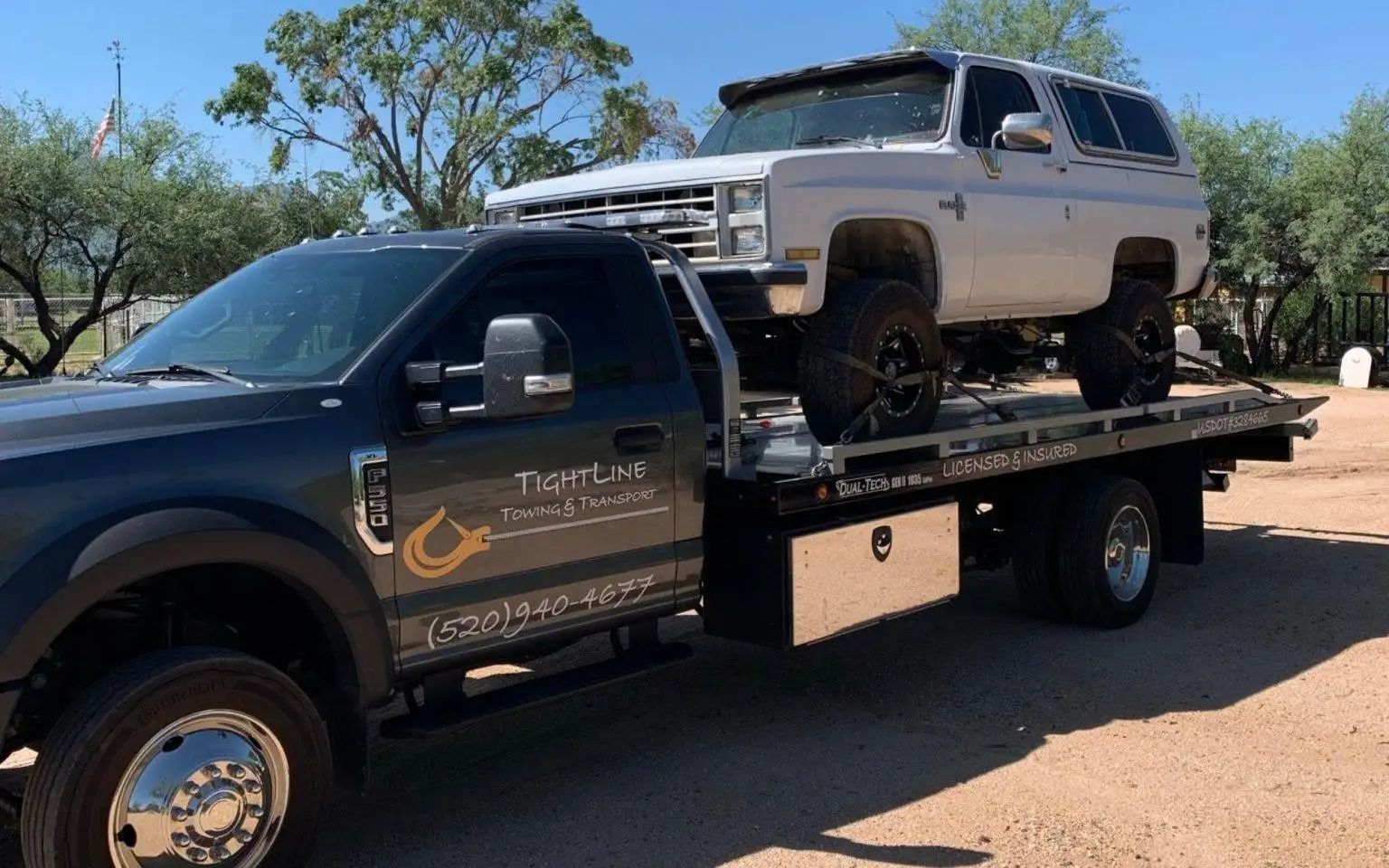 A gray pickup truck towing a lifted white SUV on a flatbed in an outdoor setting.