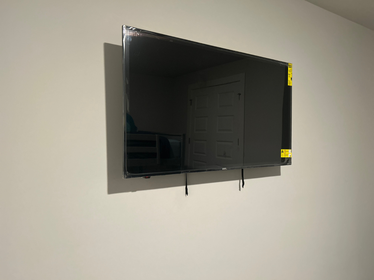 A black television mounted on a plain, light-colored wall with its protective plastic film still partially attached.