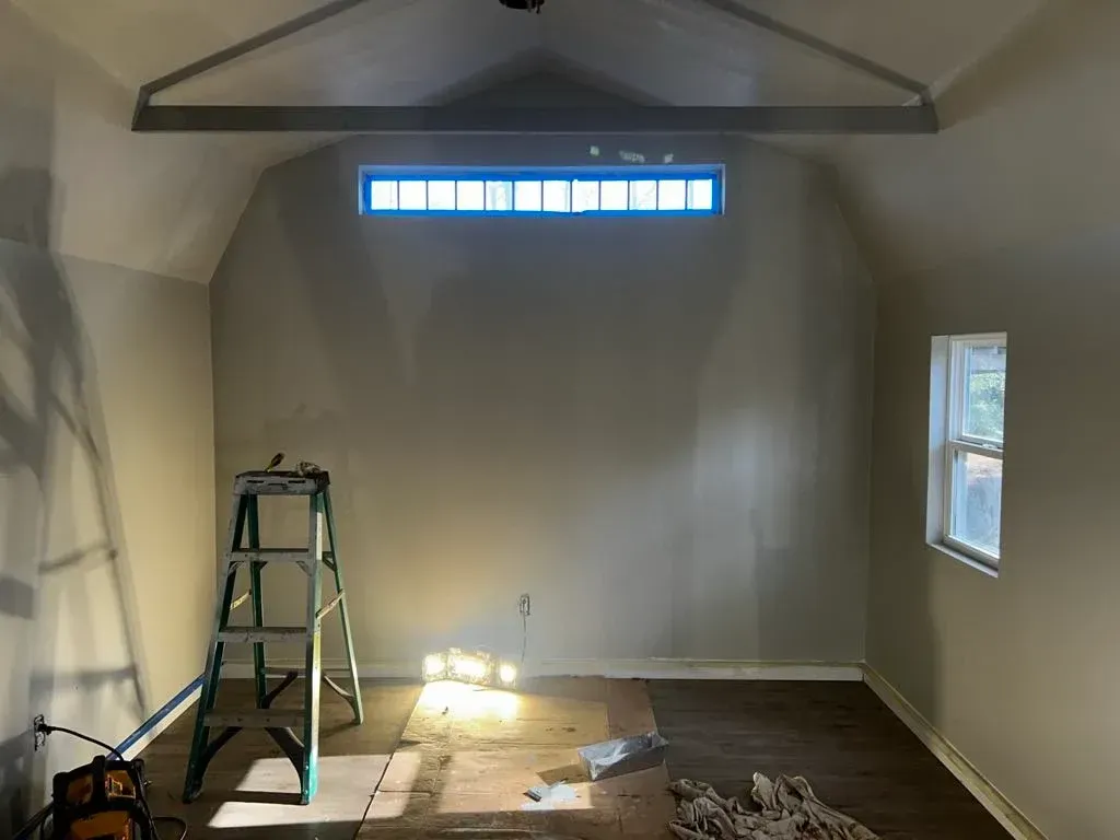 Interior view of room under construction; ladder, small window, large window, exposed beams, neutral walls.