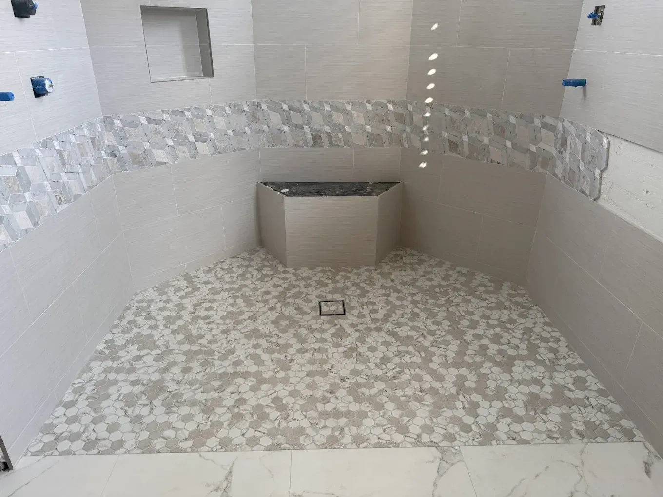 Bathroom with white and patterned tiles, angled shower floor with central drain, and small alcove.