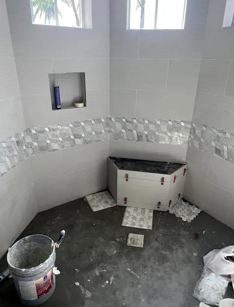 Bathroom renovation in progress; gray tile walls with accent strip, a built-in bench, and grout bucket.