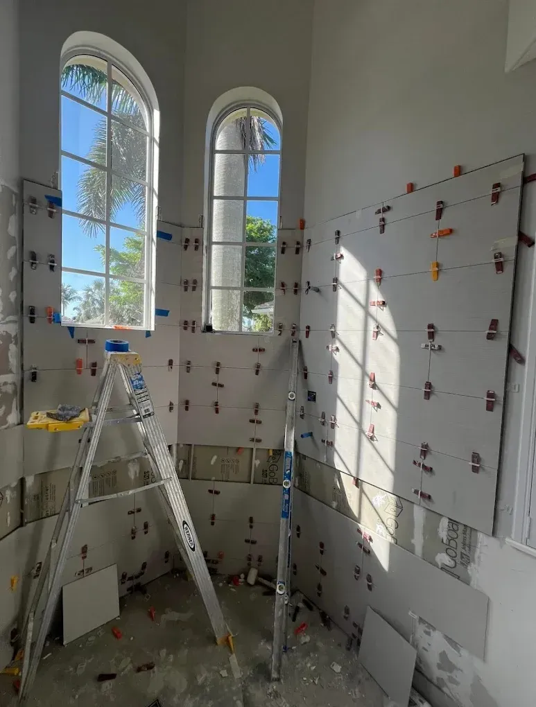 Construction: tiling a bathroom wall with large gray tiles. Tools, ladder, and windows are visible.