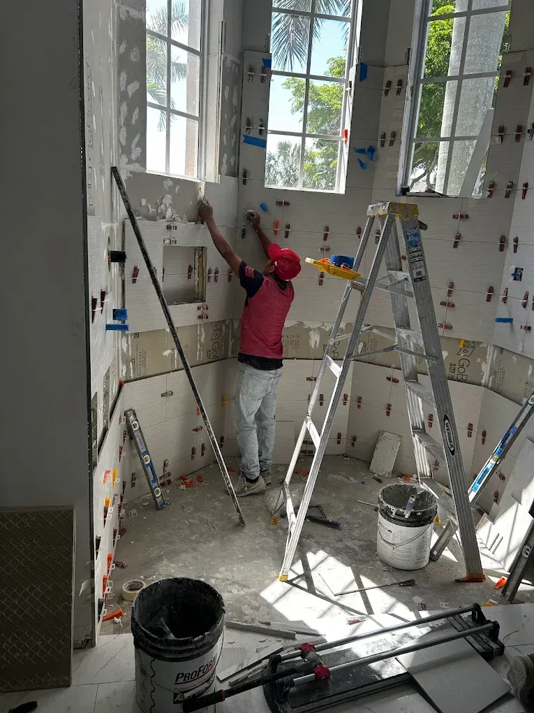 Person tiling a curved wall in a room with windows. He's using a ladder and tools.