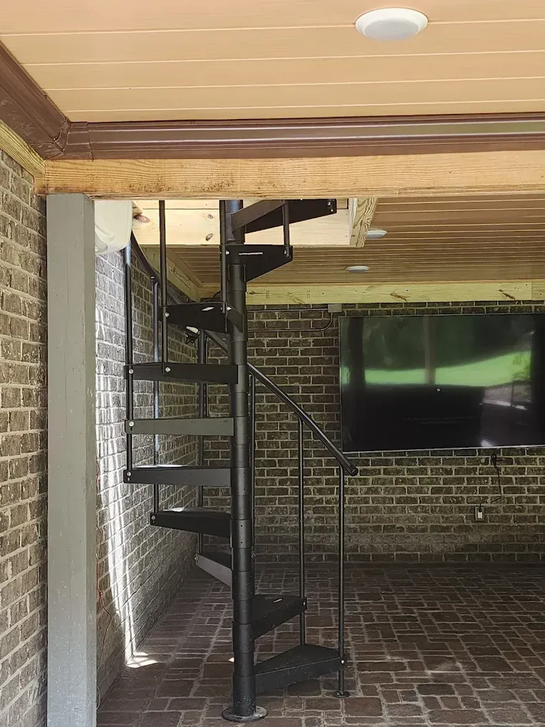 Black spiral staircase against brick walls, leading to an opening in the ceiling.
