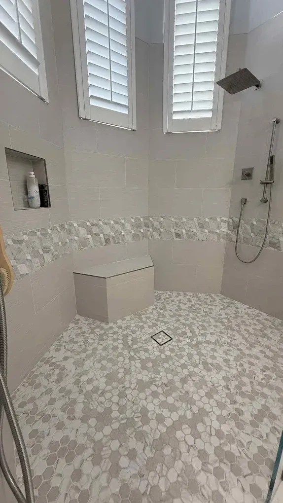 Bathroom with white and gray floral tiles, shower bench, and windows with shutters.