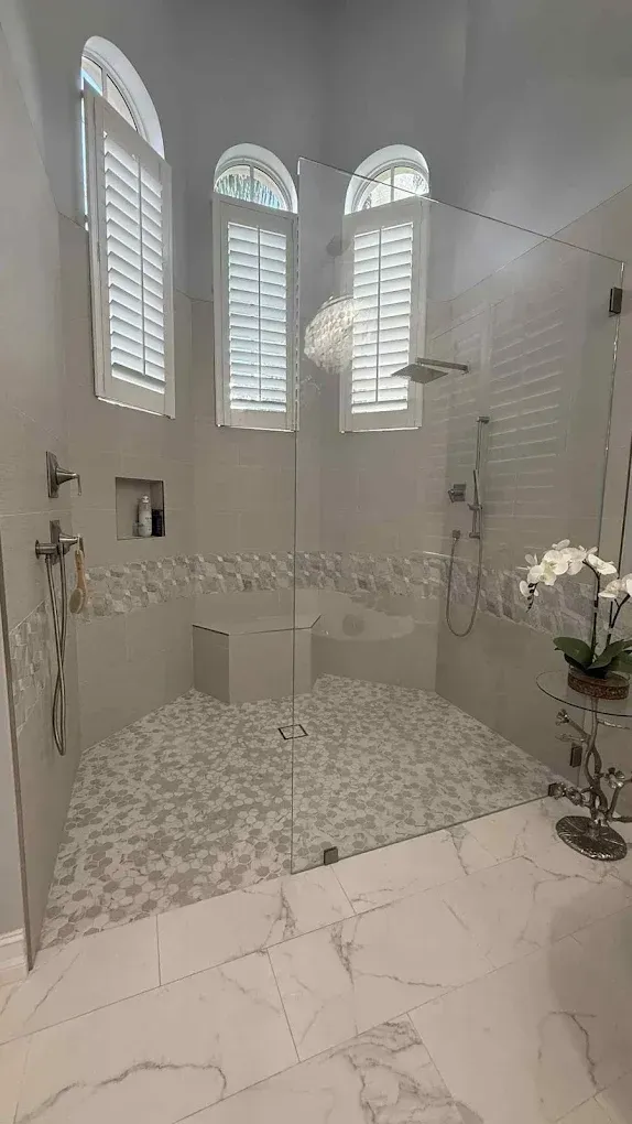 Luxurious white bathroom with glass shower, mosaic floor, arched windows with shutters, and a plant.