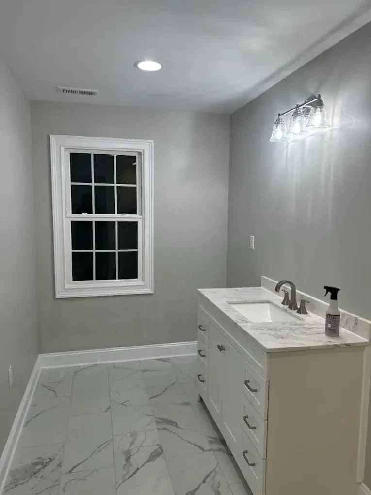A bathroom with gray walls, white vanity, and marble-look floor. A window is on the left.