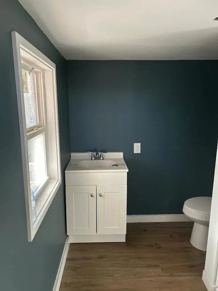 Small bathroom with white vanity, toilet, and blue walls. Window on the left.