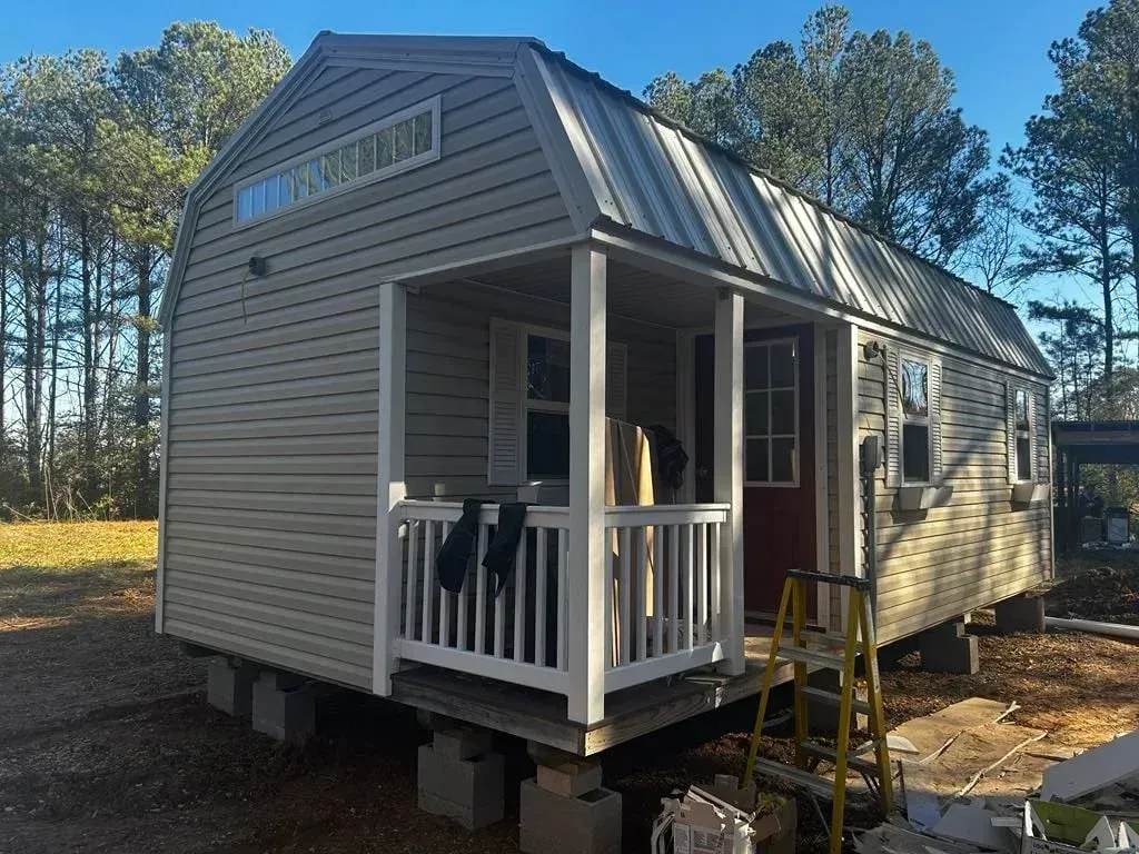 Tan-sided shed with a porch, red door, and metal roof, sitting on concrete blocks in a wooded area.