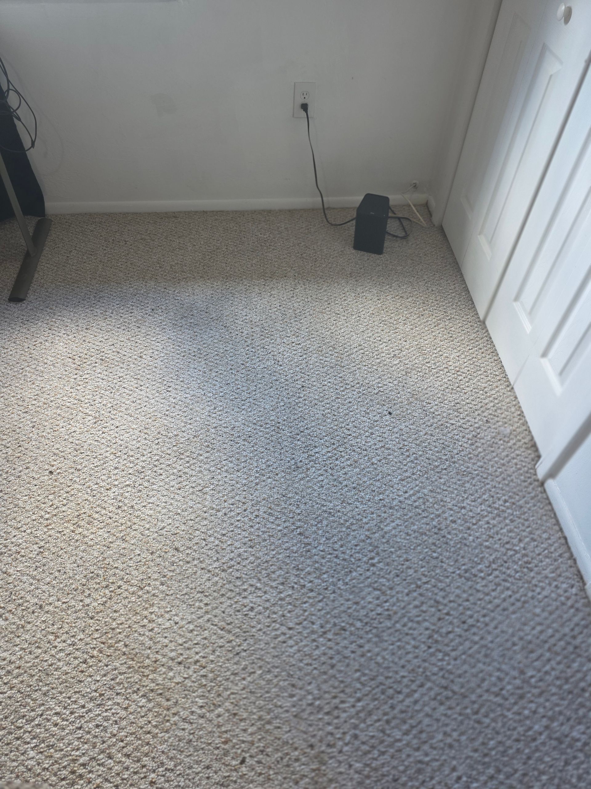 A view of a grey-flecked carpet in a corner of a room, featuring a wall outlet and a small black electronic device.