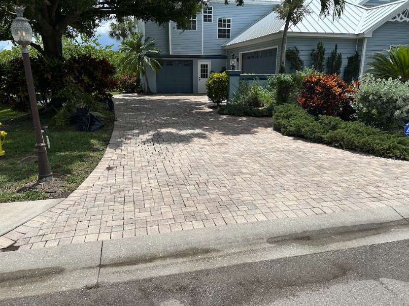 Brick driveway leading to a blue house with a two-car garage, surrounded by landscaping and trees.