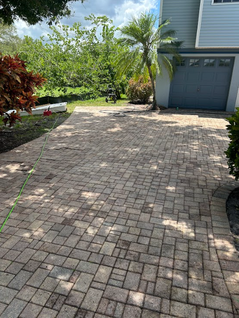 Brick driveway leading to a light blue house with a grey garage door, surrounded by greenery.