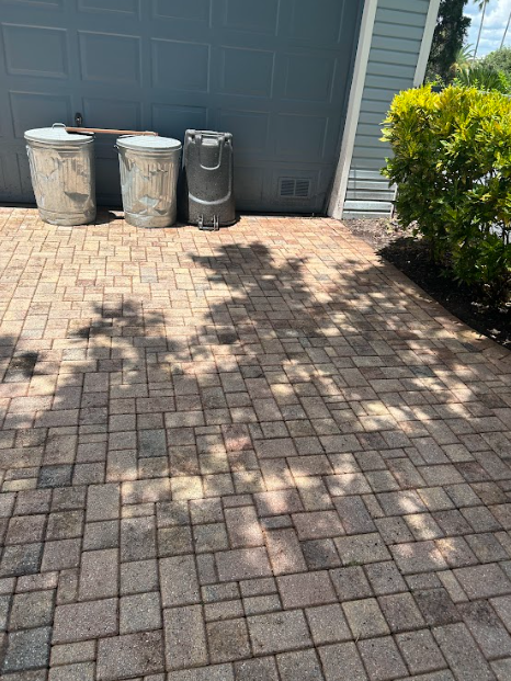 Brick patio with shadows, three trash cans against a blue garage door, next to a green bush.