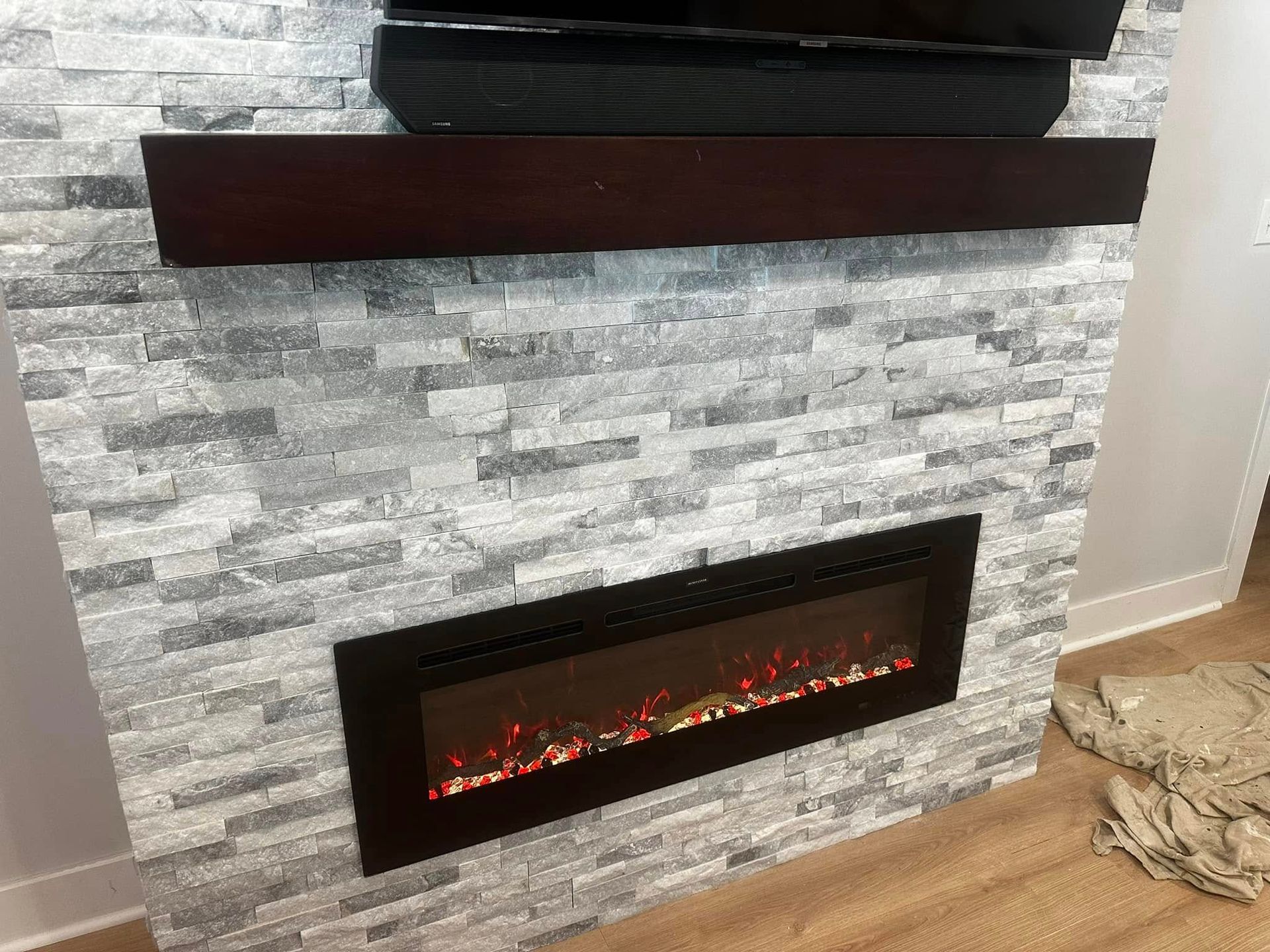 Electric fireplace with gray stone facing, below a dark wood shelf and a TV.