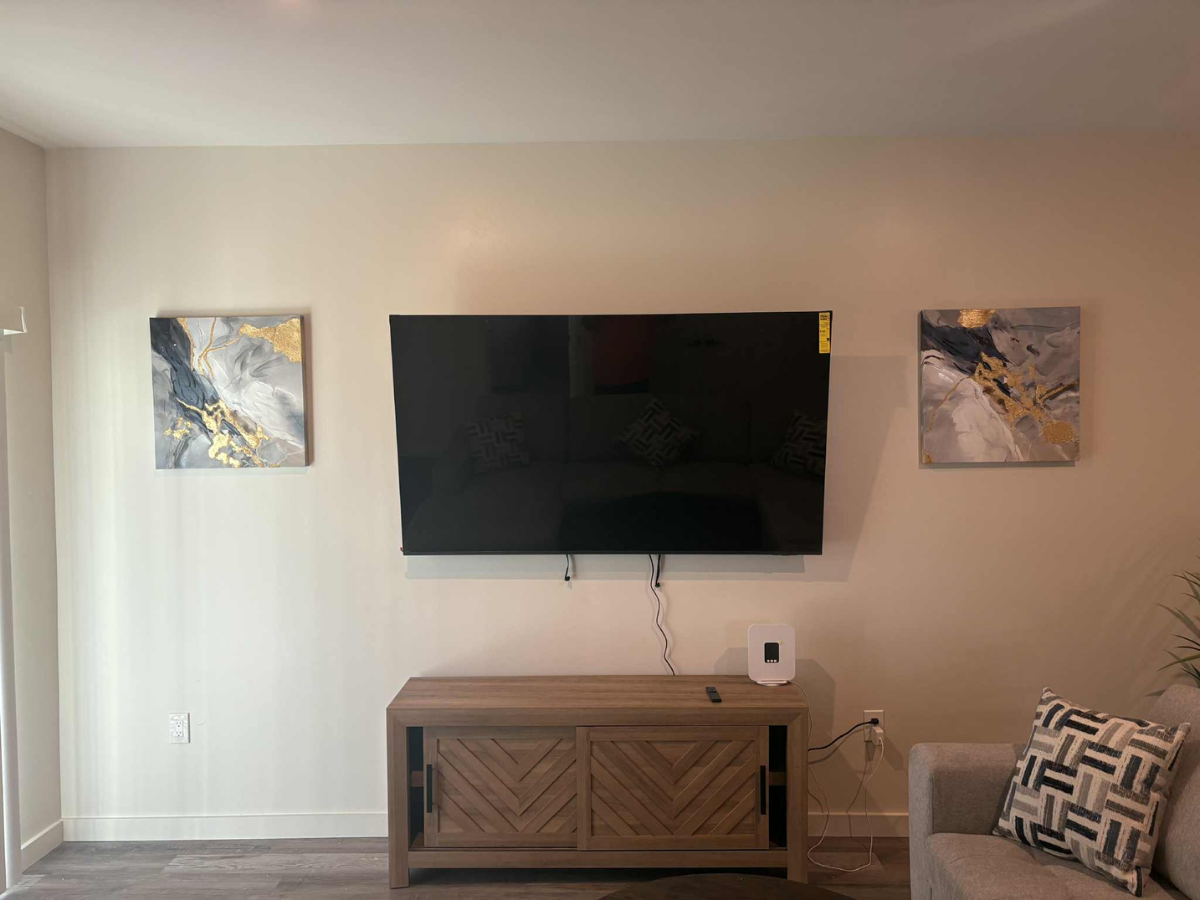 A wall-mounted TV flanked by two abstract, gold and grey canvas paintings above a light wooden media console.