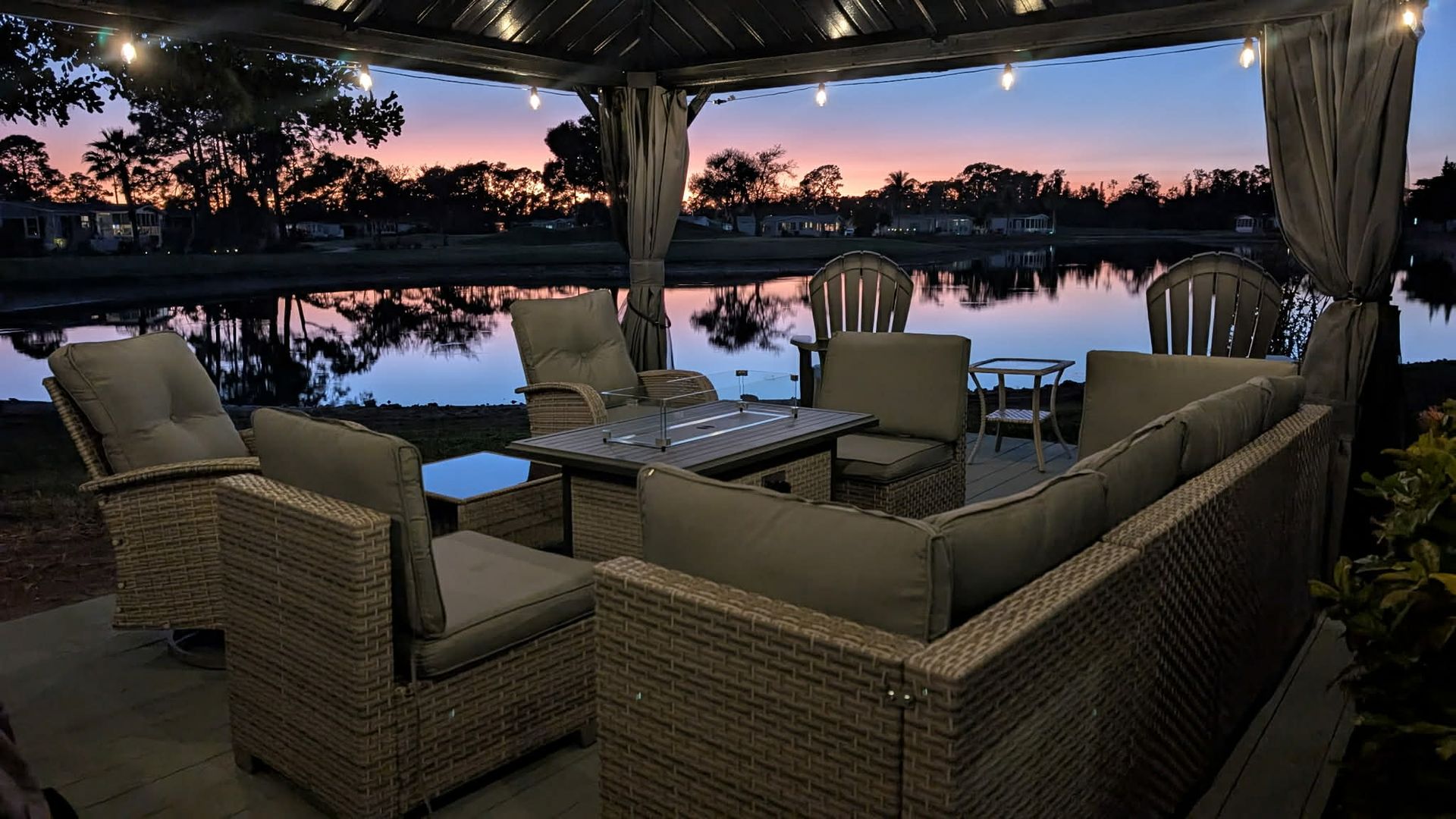 Wicker patio furniture under a lighted gazebo overlooks a calm lake at sunset.