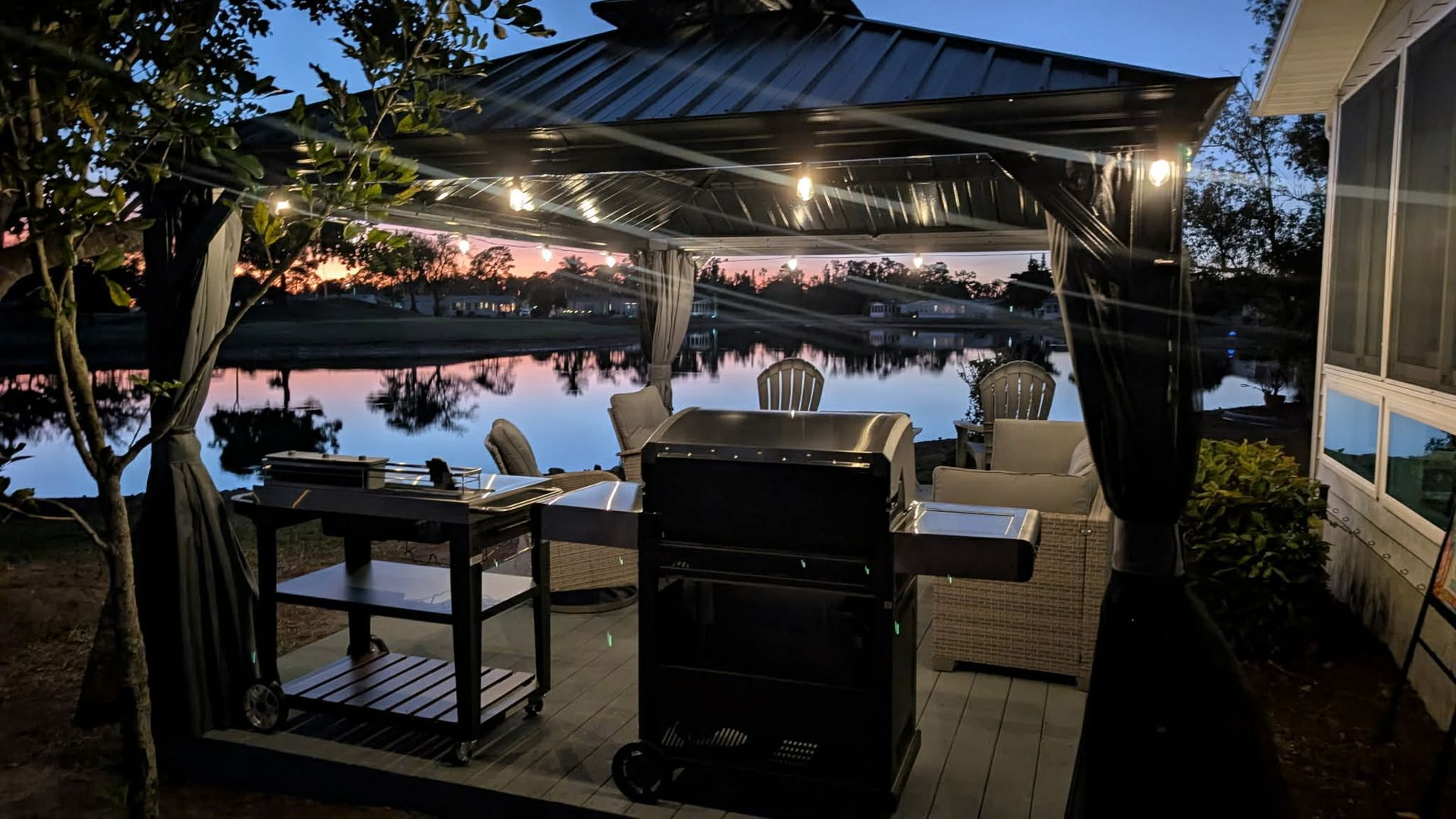 A black gazebo with string lights sits on a patio next to a grill, overlooking a lake at sunset.
