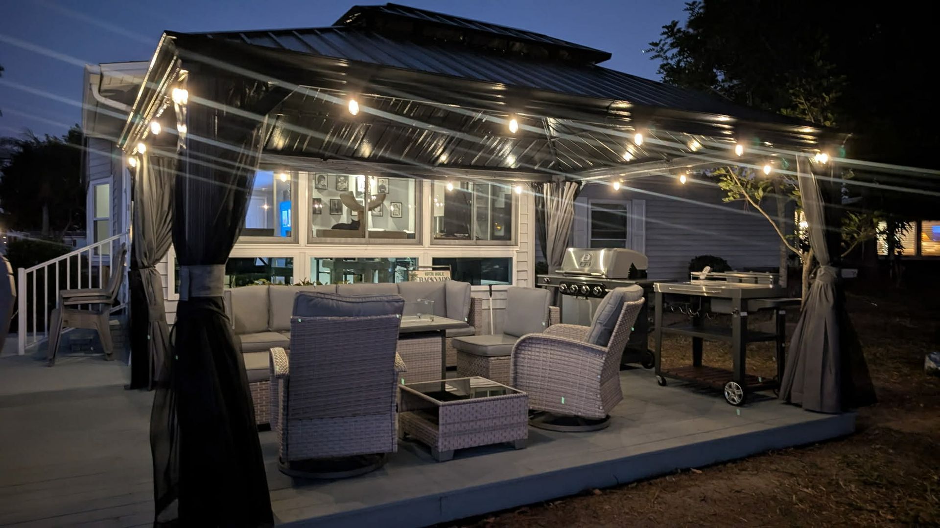 A backyard patio at dusk featuring a metal-roofed gazebo with string lights, gray outdoor furniture, and a metal grill.