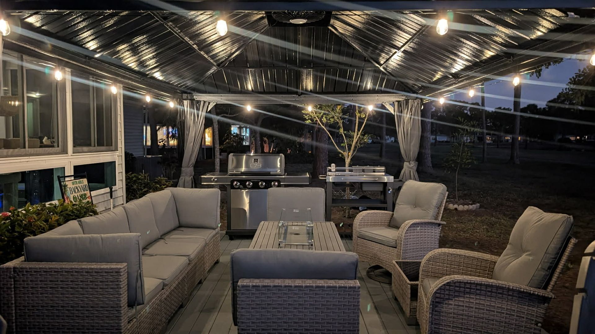 A lit outdoor patio at dusk, featuring a grey wicker sofa set, chairs, a coffee table, and a stainless steel gas grill.