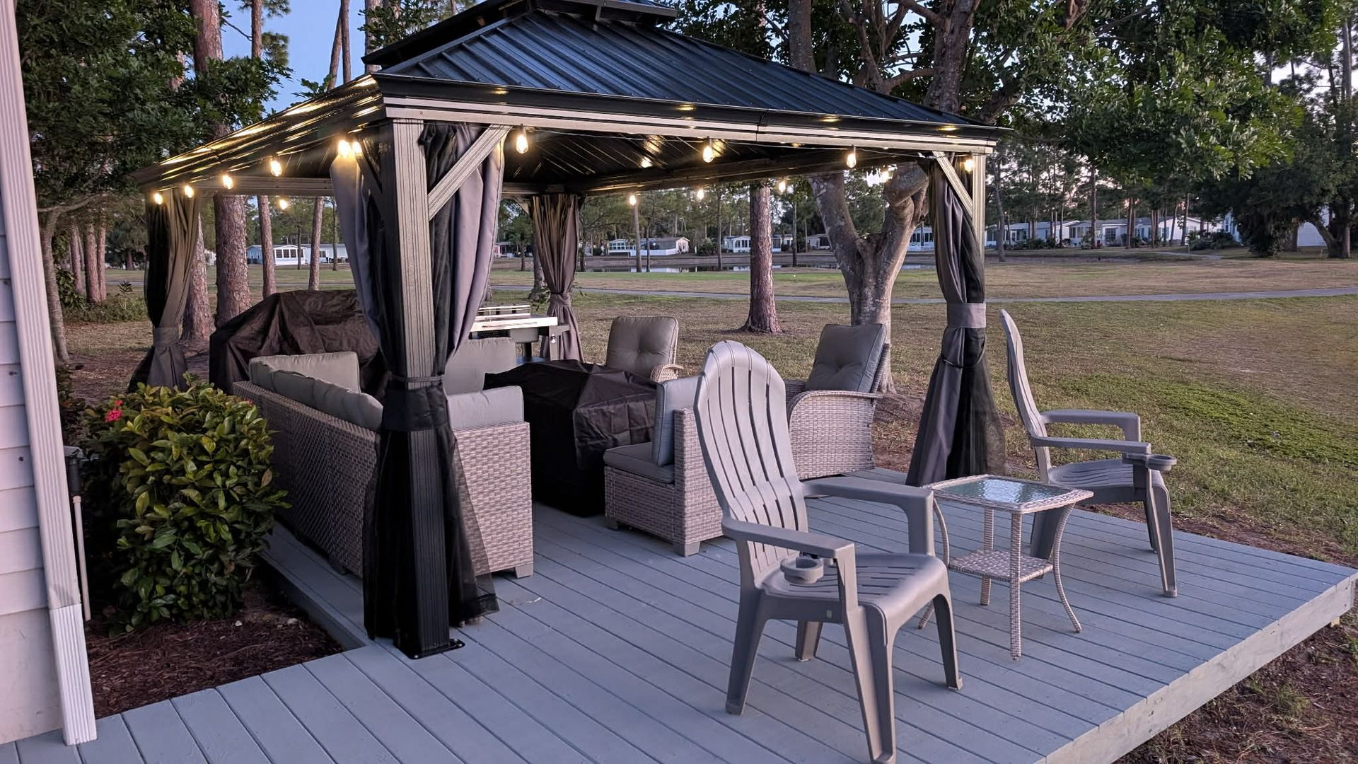 A gazebo with string lights stands on a grey deck, furnished with wicker seating and plastic chairs outdoors at dusk.