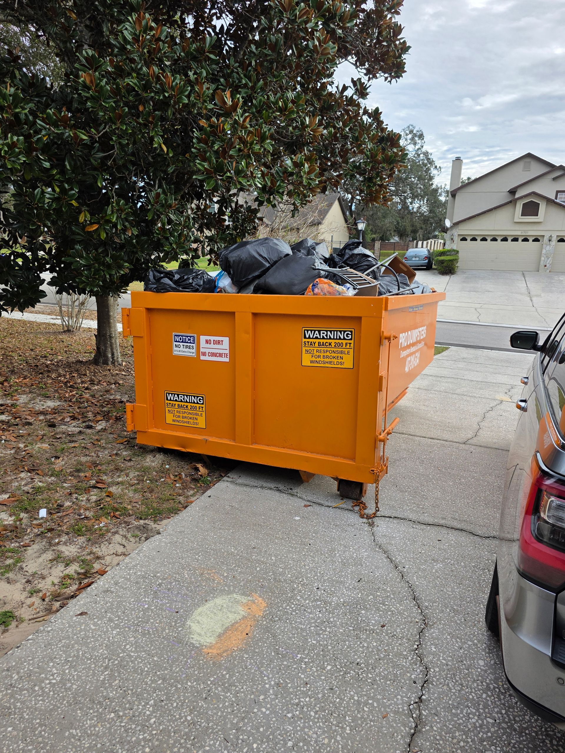 An orange dumpster filled with trash bags sits on a residential driveway next to a large tree and a parked car.