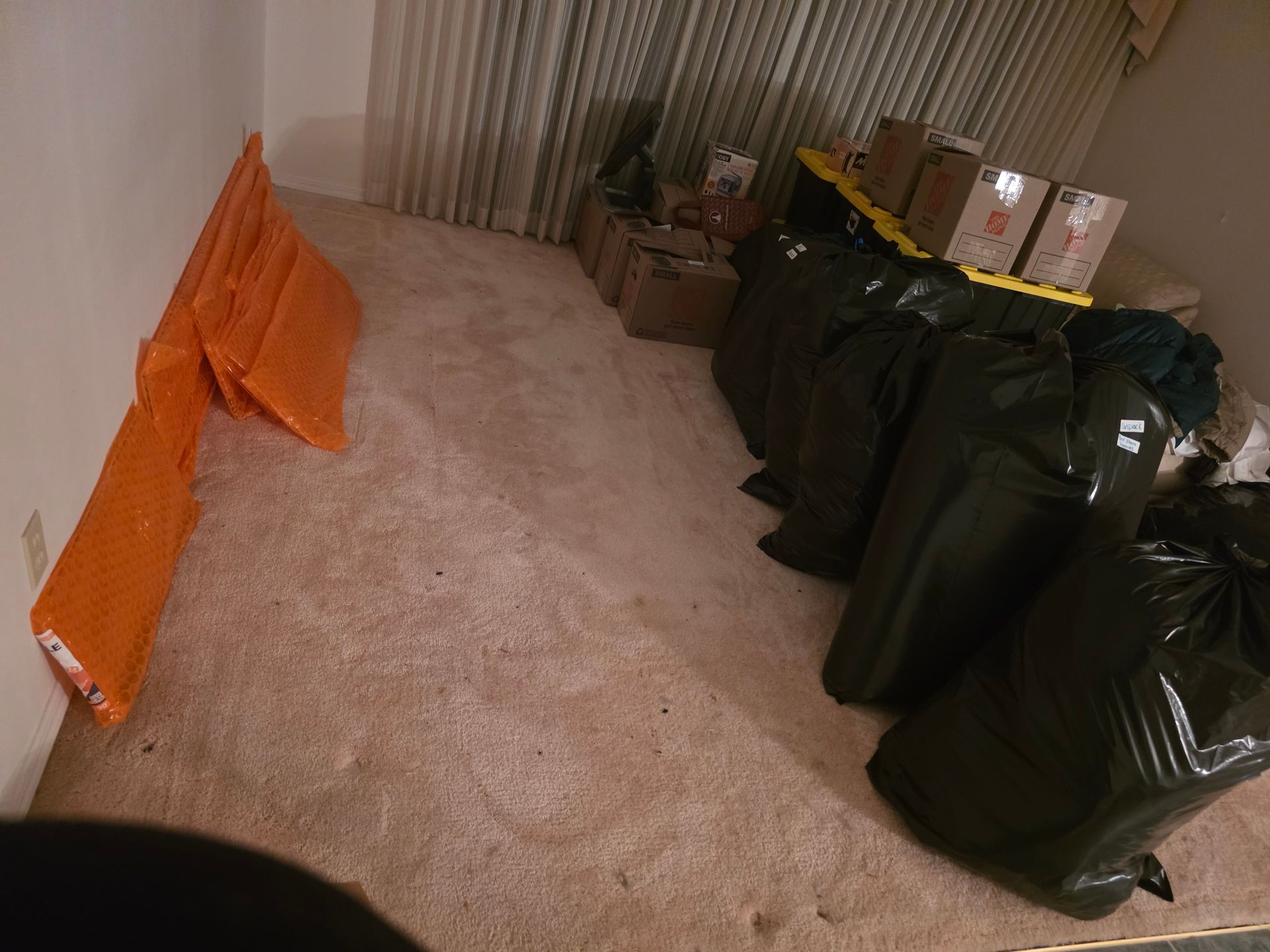 Boxes and black bags are stacked along a wall next to a vertical orange panel in a room with beige carpet.