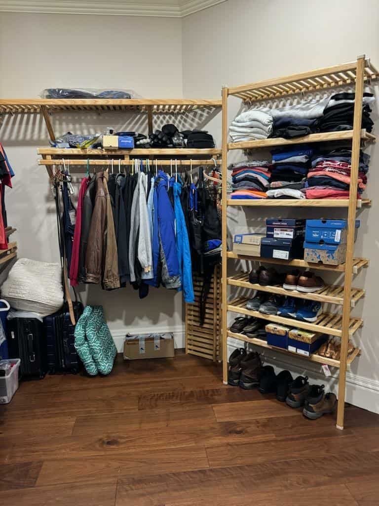 A wooden closet organizer with shelves holding folded clothes, shoes, and hanging jackets against a neutral wall.