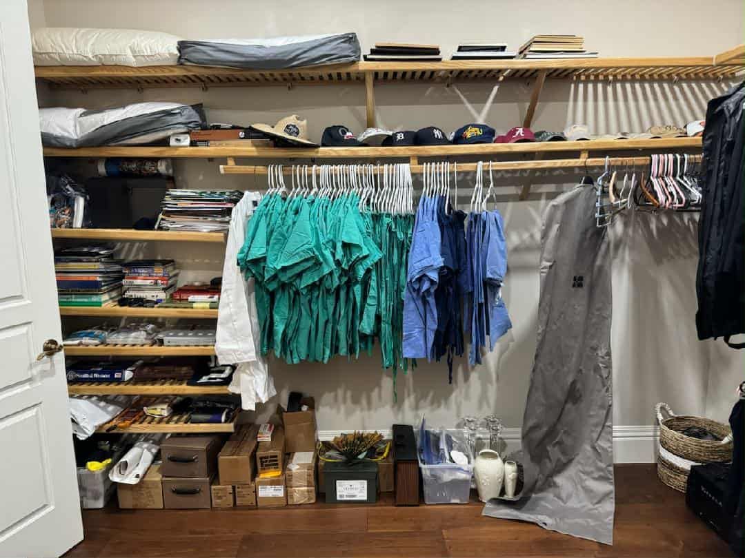 A walk-in closet with shelves holding folded items, hanging medical scrubs in green and blue, and boxes on the floor.