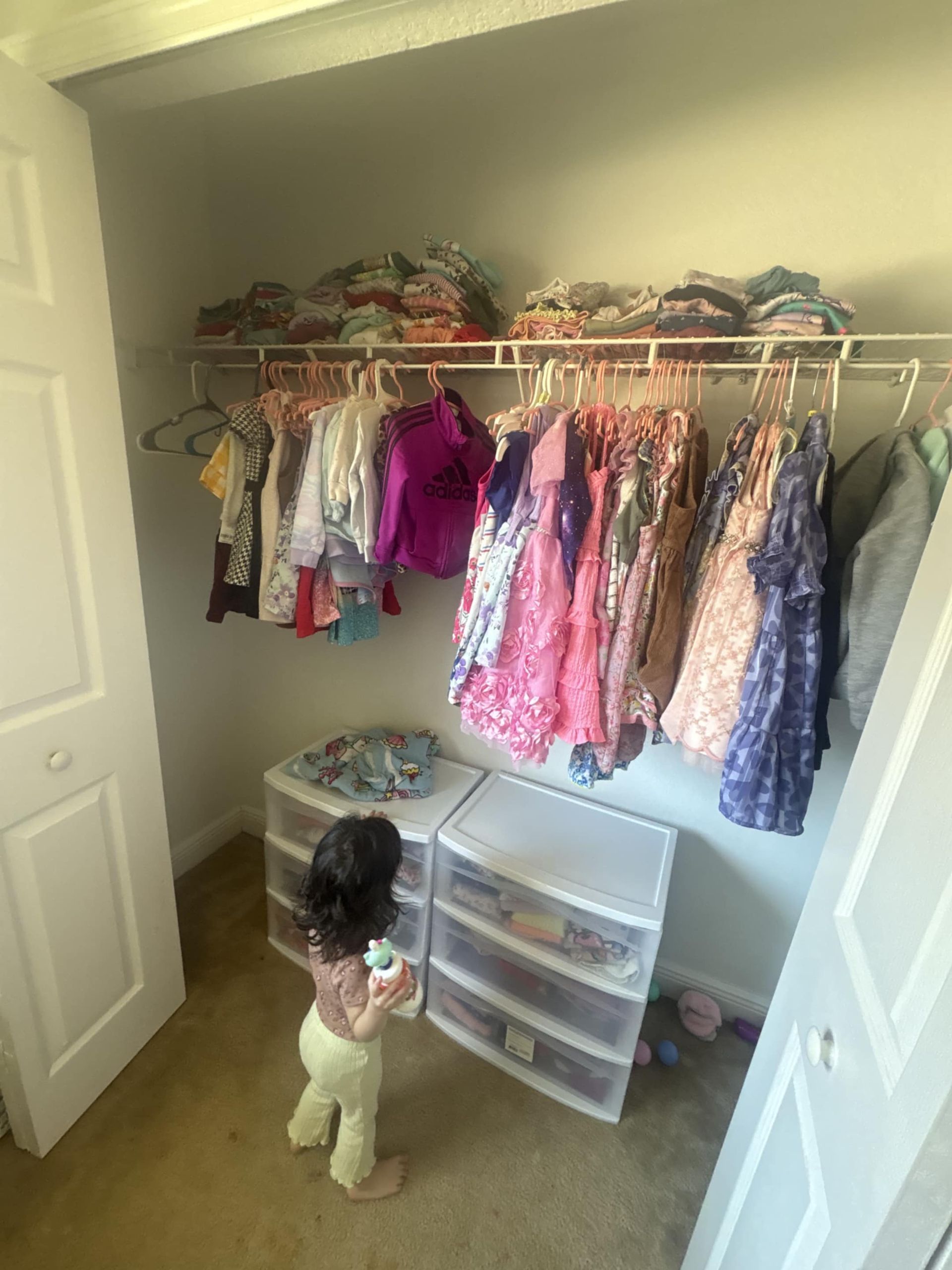 A child standing in a closet facing two stacked plastic drawer units beneath a rack of hanging clothing.