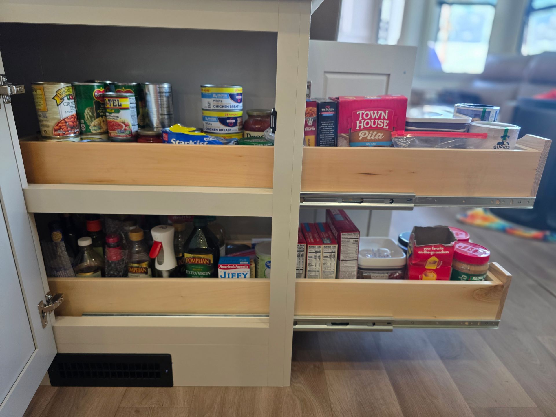 A kitchen cabinet with two pull-out wooden shelves filled with canned goods, boxed food, and various pantry items.