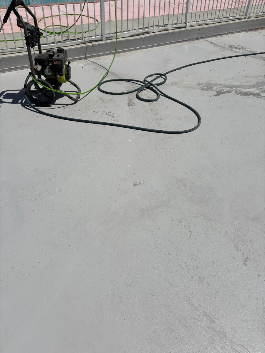 A pressure washer sits on a light gray concrete patio near a white railing, with a black hose coiled on the ground.